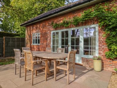 An outdoor patio with a wooden dining table and six chairs next to a brick house with glass doors and windows at Rookery Cottage in Kelsale near Saxmundham
