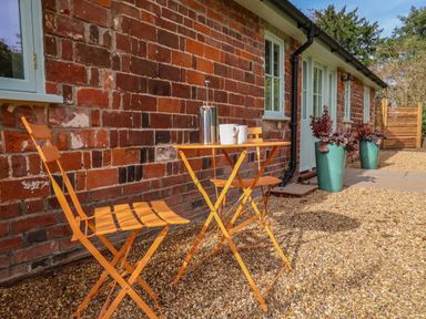A small outdoor seating area with an orange table and two orange chairs next to a brick building with windows and two large green plant pots in front at Rookery Cottage in Kelsale near Saxmundham