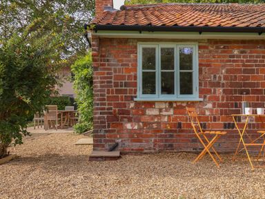 A brick cottage wall with a blue window and orange garden chairs and table on gravel at Rookery Cottage in Kelsale near Saxmundham