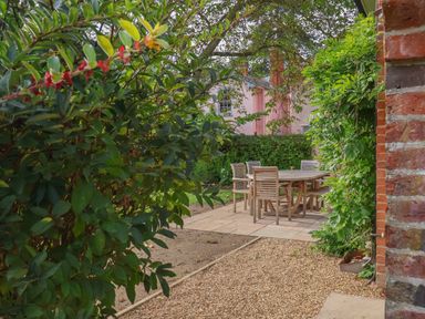 An outdoor patio area with wooden table and chairs surrounded by green plants and gravel path at Rookery Cottage in Kelsale near Saxmundham