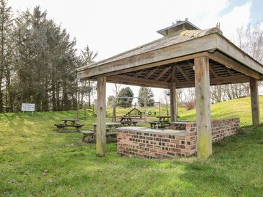 An outdoor covered seating area with wooden picnic tables a brick structure and playground equipment in a grassy area at Matfen in Longframlington