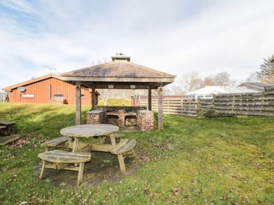 A wooden picnic table and benches on a grassy area near a covered brick barbecue shelter with wooden posts and a sloped roof at Matfen in Longframlington