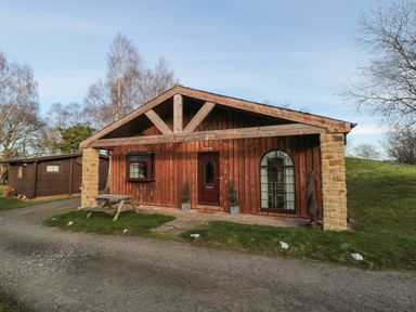 A wooden cabin with a picnic table outside on a lawn near a gravel driveway at Matfen in Longframlington