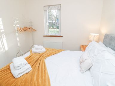 A bedroom with a neatly made bed with white and mustard bedding folded towels on the bed a window with a blind a bedside table with a lamp and a clothes rack with hangers
