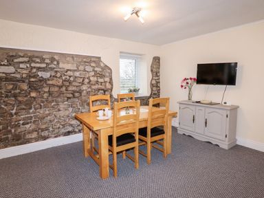 A dining room with a wooden table and chairs a stone accent wall a window a wall mounted TV and a cabinet with flowers at Fern Cottage in St Brides-Super-Ely