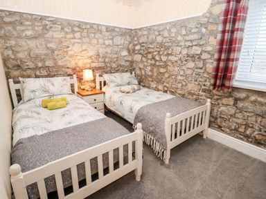 A bedroom with two single beds a nightstand with a lamp stone walls carpeted floor and a window with red plaid curtains at Fern Cottage in St Brides-Super-Ely