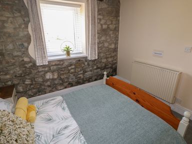 A bedroom with a bed covered with a blue blanket and tropical print sheets stone wall with a window and curtains and a radiator on the right wall at Fern Cottage in St Brides-Super-Ely