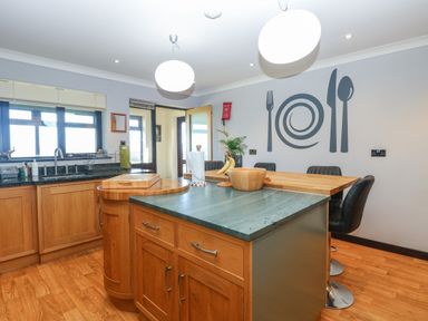 A kitchen with wooden cabinets an island with a countertop four bar stools and wall art of a fork plate knife and spoon at Beach Haven in Happisburgh East Anglia