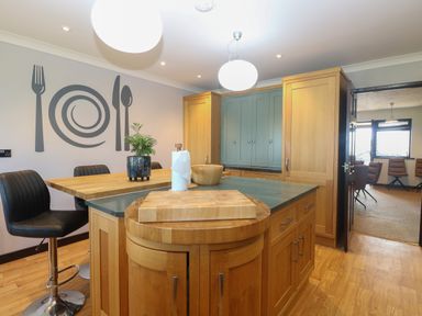 A kitchen with wooden cabinets a countertop with a cutting board a dining table with chairs and wall art of utensils at Beach Haven in Happisburgh East Anglia