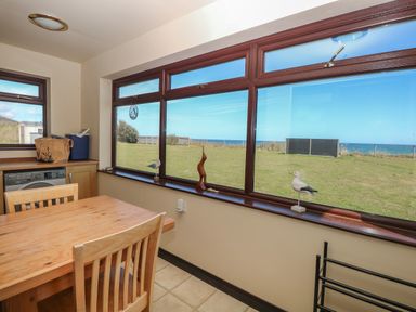 A dining area with a wooden table and chairs next to large windows overlooking a grassy yard and ocean at Beach Haven in Happisburgh East Anglia