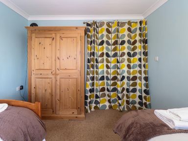 A bedroom with a wooden wardrobe two beds with brown covers and colorful patterned curtains at Beach Haven in Happisburgh East Anglia
