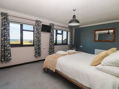 A bedroom with a bed white linens and beige pillows two windows with patterned curtains and a wall-mounted tv at Beach Haven in Happisburgh East Anglia