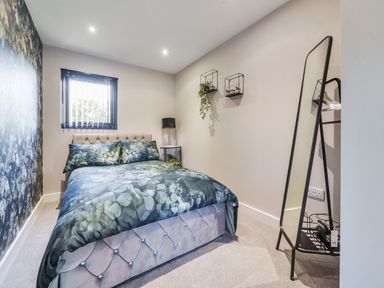 A bedroom with a bed with floral bedding a window with blinds wall shelves with plants a floor mirror and a carpeted floor at Micklemore Lakes and Lodges in North Thoresby