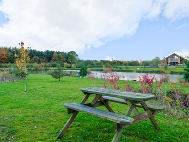 A wooden picnic table on grass near a pond with trees and a house in the background at Micklemore Lakes and Lodges North Thoresby
