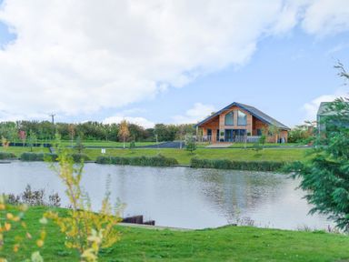 A wooden house near a lake with trees and grass around at Micklemore Lakes and Lodges in North Thoresby