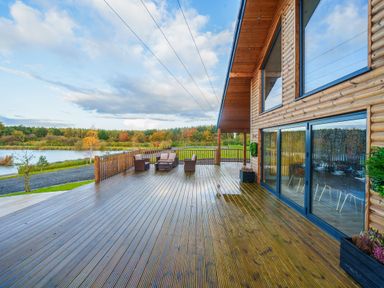 A large wooden deck with outdoor seating next to a log cabin overlooking a lake and trees at Micklemore Lakes and Lodges in North Thoresby