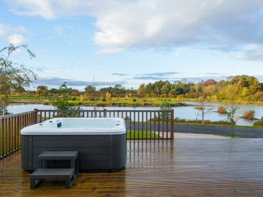 An outdoor hot tub on a wooden deck overlooking a lake with trees and shrubs at Micklemore Lakes and Lodges in North Thoresby