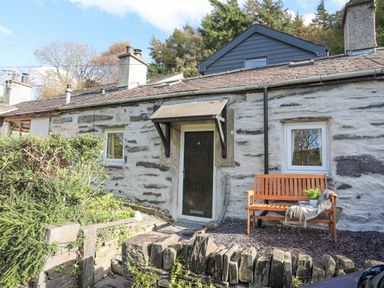 The exterior of a stone cottage with a wooden door a wooden bench with a plant and cloth next to the door and greenery around the entrance at 4 Ty Person in Braichmelyn near Bethesda