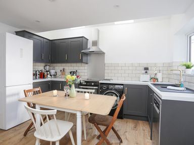 A kitchen with dark cabinets a wooden table with chairs and appliances at 4 Ty Person in Braichmelyn near Bethesda