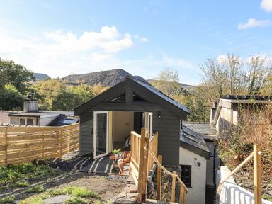 An outdoor view of a small dark gray building with open doors leading to an interior room a wooden fence and steps with rope railing at 4 Ty Person in Braichmelyn near Bethesda
