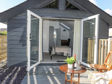 A small house with open glass doors showing a bedroom inside and a wooden patio with a table and chairs at 4 Ty Person in Braichmelyn near Bethesda