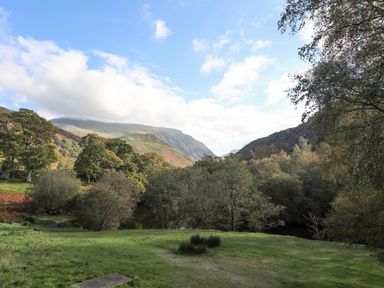 A grassy field with trees and hills under a blue sky at 4 Ty Person in Braichmelyn near Bethesda