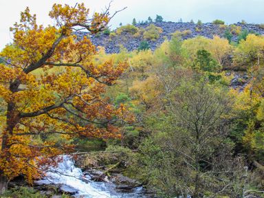 A river with a small waterfall surrounded by autumn trees and a rocky hill in the background at 4 Ty Person Braichmelyn near Bethesda