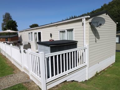 An exterior view of a beige mobile home with white railing around a porch and a satellite dish on the side at 116 Woodpecker Lodge in Gilfachrheda near Llanarth