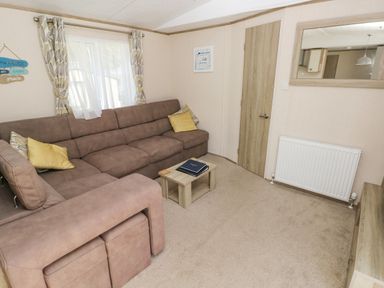 A living room with a brown sectional sofa yellow pillows a small wooden coffee table a window with curtains a door a radiator and a wall mirror at 116 Woodpecker Lodge in Gilfachrheda near Llanarth