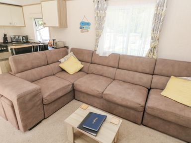 A living room with a brown sectional sofa yellow pillows a small wooden coffee table and a kitchen area at 116 Woodpecker Lodge in Gilfachrheda near Llanarth