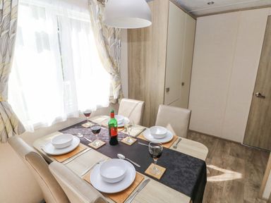 A dining table set for four with plates bowls cutlery wine glasses and a bottle in a room with curtains and wooden floor at 116 Woodpecker Lodge in Gilfachrheda near Llanarth