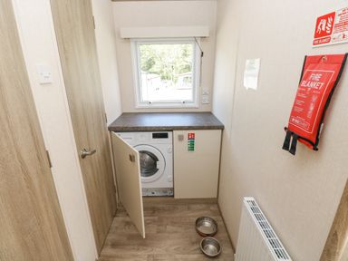 A laundry room with a washing machine with open cabinet under a window and fire blanket on wall at 116 Woodpecker Lodge in Gilfachrheda near Llanarth