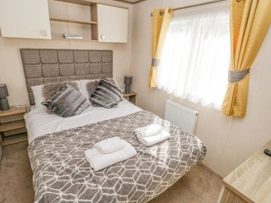 A bedroom with a double bed pillows bedside tables lamps window with yellow curtains and folded towels on the bed at 116 Woodpecker Lodge in Gilfachrheda near Llanarth