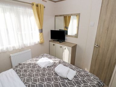 A bedroom with a bed covered with a patterned blanket and towels a cabinet with a television and a mirror above at 116 Woodpecker Lodge in Gilfachrheda near Llanarth