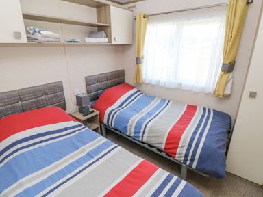 A bedroom with two single beds with striped bedcovers a nightstand a lamp shelves with towels and a window with yellow curtains at 116 Woodpecker Lodge in Gilfachrheda near Llanarth