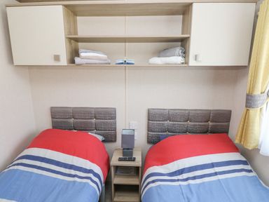 A bedroom with two single beds with red blue and white bedding and a small wooden nightstand between them at 116 Woodpecker Lodge in Gilfachrheda near Llanarth
