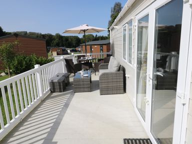 An outdoor patio with wicker furniture including chairs and tables a white fence and umbrella at 116 Woodpecker Lodge in Gilfachrheda near Llanarth