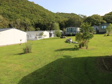 A grassy area with several small trees and bushes with holiday lodges and wooded hills in the background at 116 Woodpecker Lodge in Gilfachrheda near Llanarth