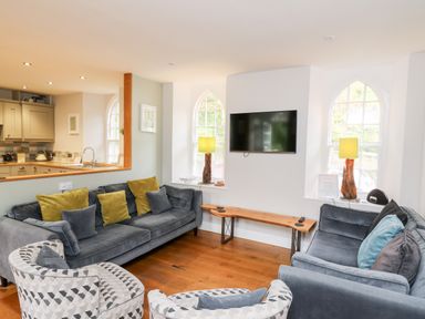A living room with sofas, patterned armchairs, a wall-mounted TV, wooden bench, and lamps by windows at Upper Vestry in Llangrannog