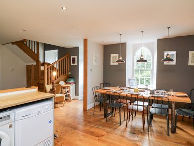 A dining area with a wooden table set with plates and glasses and metal chairs near a window and wooden stairs at Upper Vestry in Llangrannog