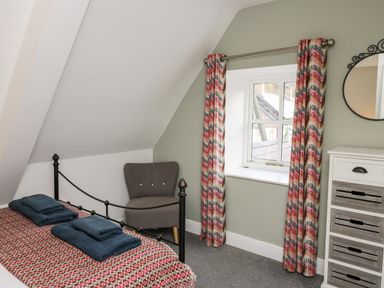 A small bedroom with a bed a gray chair a window with patterned curtains and a chest of drawers at Upper Vestry in Llangrannog
