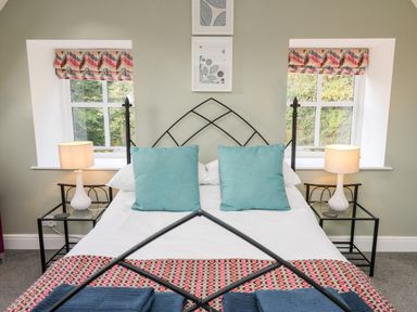 A bedroom with a metal framed bed with blue pillows bedside tables with lamps and two windows with patterned curtains at Upper Vestry in Llangrannog