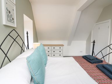 A bedroom with a bed with blue pillows and a patterned blanket a dresser and a small door under a slanted ceiling at Upper Vestry in Llangrannog