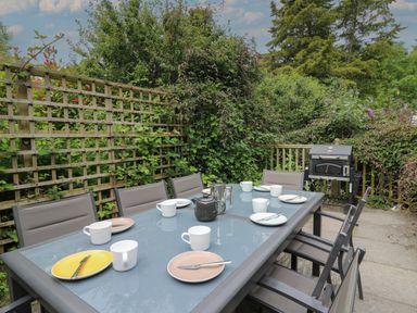 An outdoor patio with a glass table set with cups and plates surrounded by chairs and a barbecue grill at Upper Vestry in Llangrannog