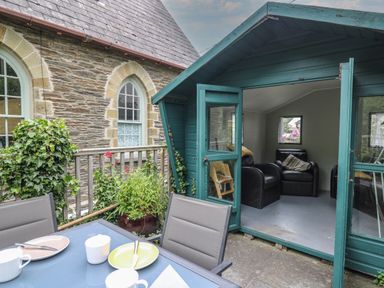 An outdoor seating area with a table and chairs next to a small green wooden cabin with armchairs inside at Upper Vestry in Llangrannog