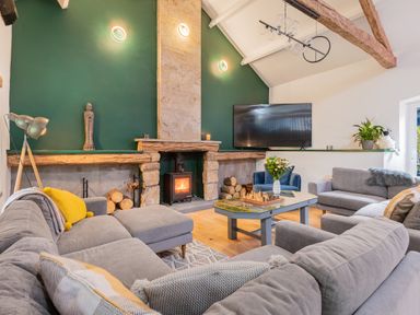 A living room with a stone fireplace a wood burning stove a large grey sectional sofa a coffee table with a chess set and flowers and a wall mounted television at The Waterfall Retreat in Orton