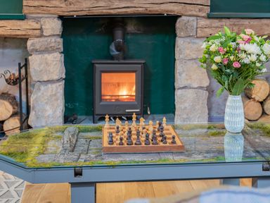 A chessboard on a glass coffee table with a vase of flowers and a wood stove in a stone fireplace at The Waterfall Retreat in Orton