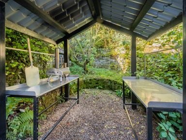An outdoor shelter with a pizza oven and metal tables surrounded by greenery and stone walls at The Waterfall Retreat in Orton