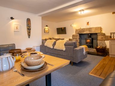 A living room with a dining table set with bowls cups and a teapot a grey sofa with throws a stone fireplace with a wood stove and a wall mounted TV at The Waterfall Retreat in Orton