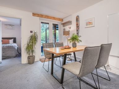A dining room with a wooden table and four grey chairs near a plant and a doorway showing a bedroom at The Waterfall Retreat in Orton
